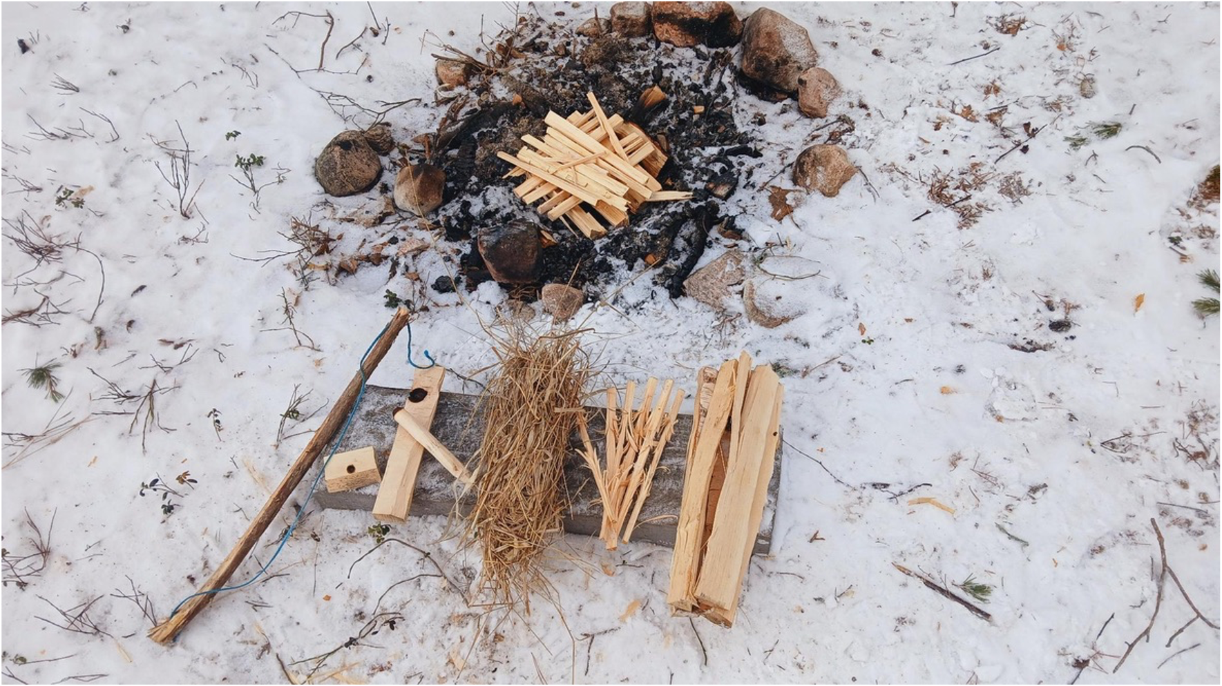 Snowy ground around a small campfire ring with ashes and rocks, plus bundles of kindling and wood nearby.